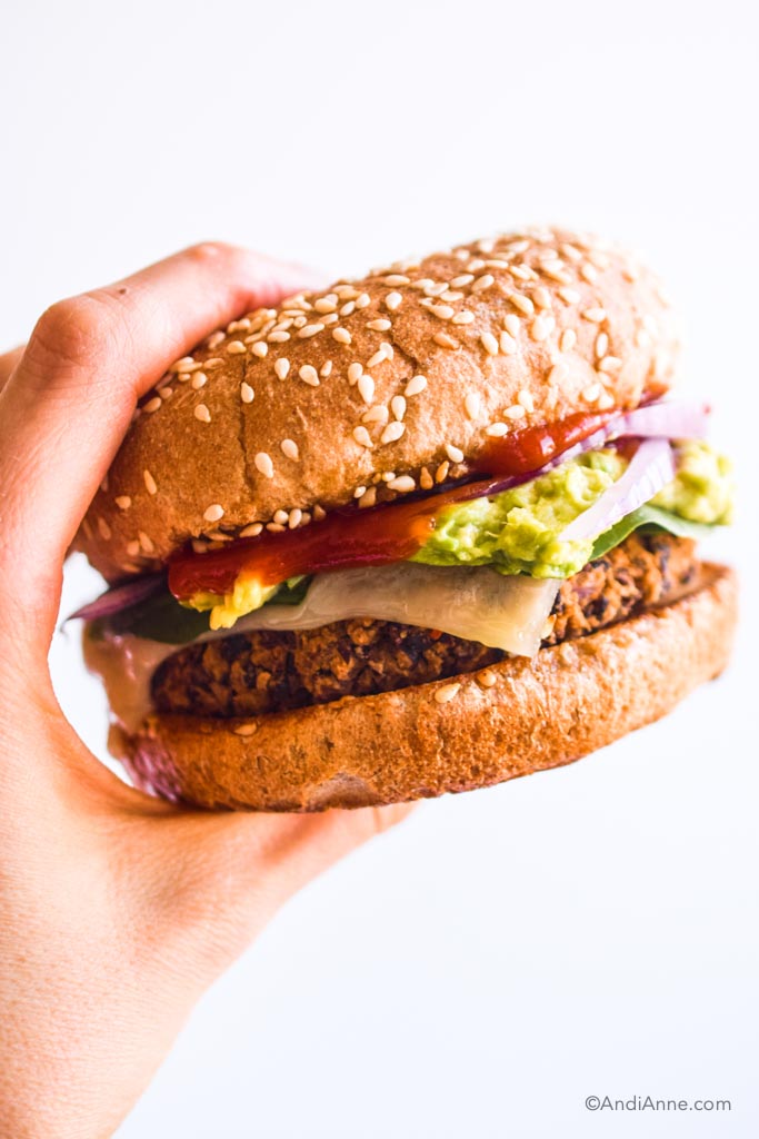 hand holding a black bean burger with ketchup, avocado, sliced onion and melted cheese