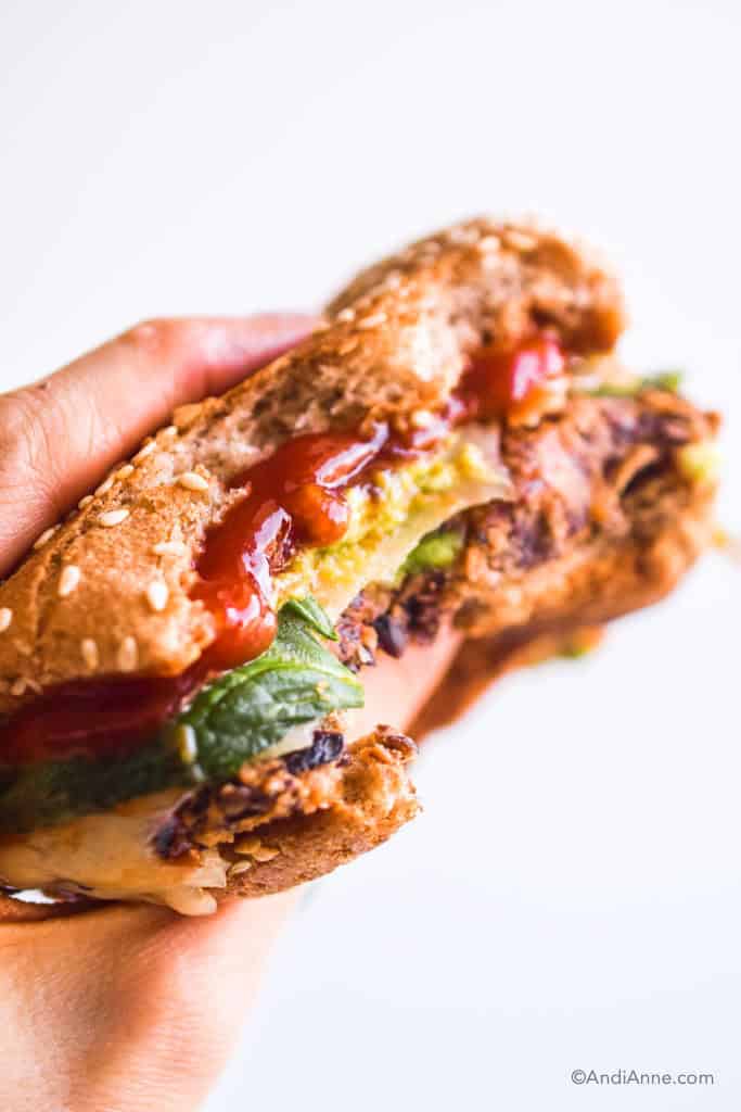 hand holding burger. big bites taken out of the black bean burger with ketchup, mashed avocado and spinach poking out.
