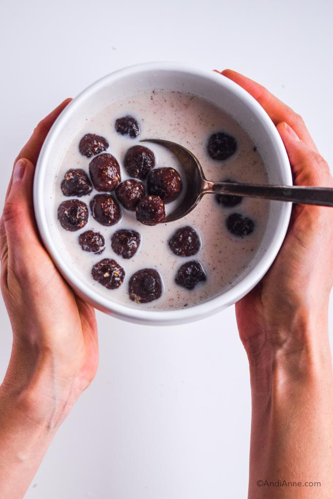 homemade chocolate cereal balls with milk in a white bowl with spoon held in hands