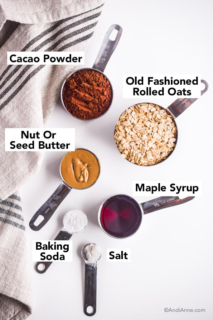 ingredients in measuring cups on a white table with kitchen towel. Cacao powder, rolled oats, nut butter, maple syrup, baking soda and salt.