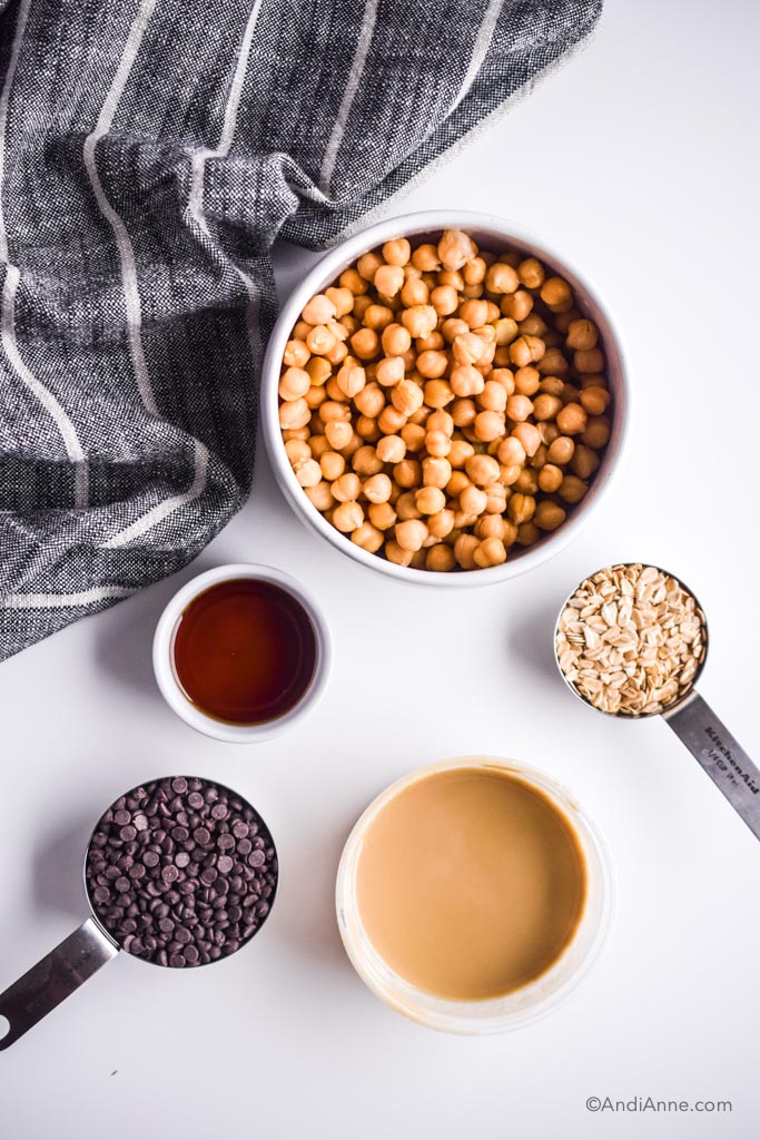 Bowl of chickpeas, and cups of rolled oats, maple syrup, tahini and chocolate chips on a white table with black and white kitchen napkin.