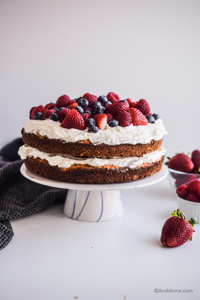 gluten free cake with fresh berries and whipped cream on a cake stand. Berries in a bowl on the side.