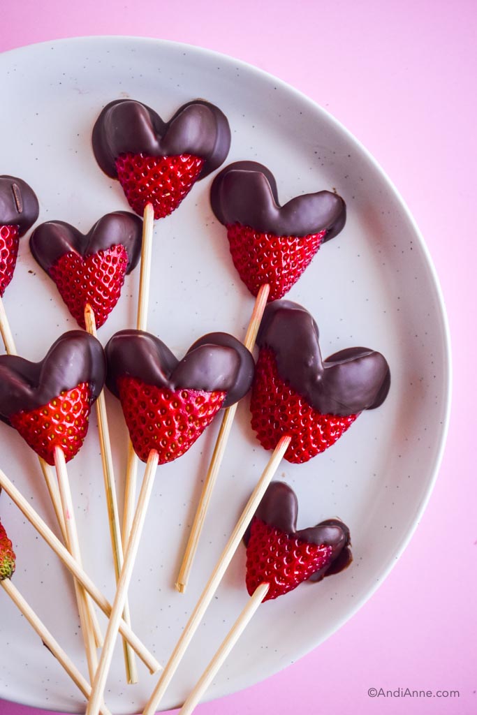 chocolate dipped heart strawberries poked with wooden skewers on a white plate with a pink background
