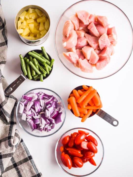 bowls of raw chicken, green beans, carrots, red onion, and sliced grape tomatoes on a white table