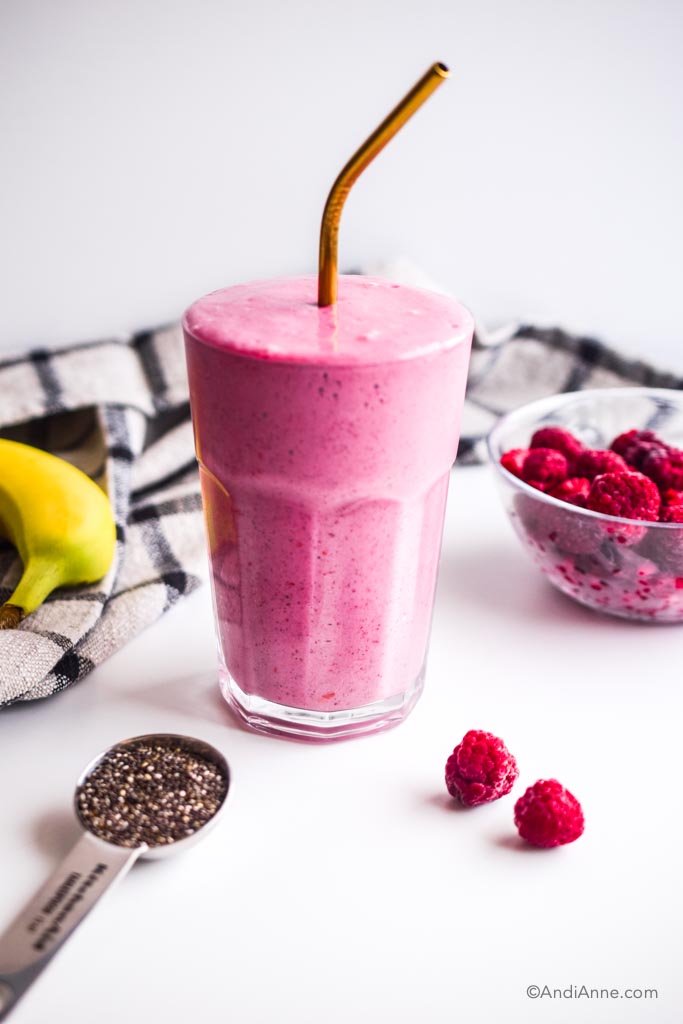 glass with raspberry yogurt smoothie inside and gold straw. Bowl of raspberries, a banana, spoon of chia seeds and a kitchen napkin surround the glass.