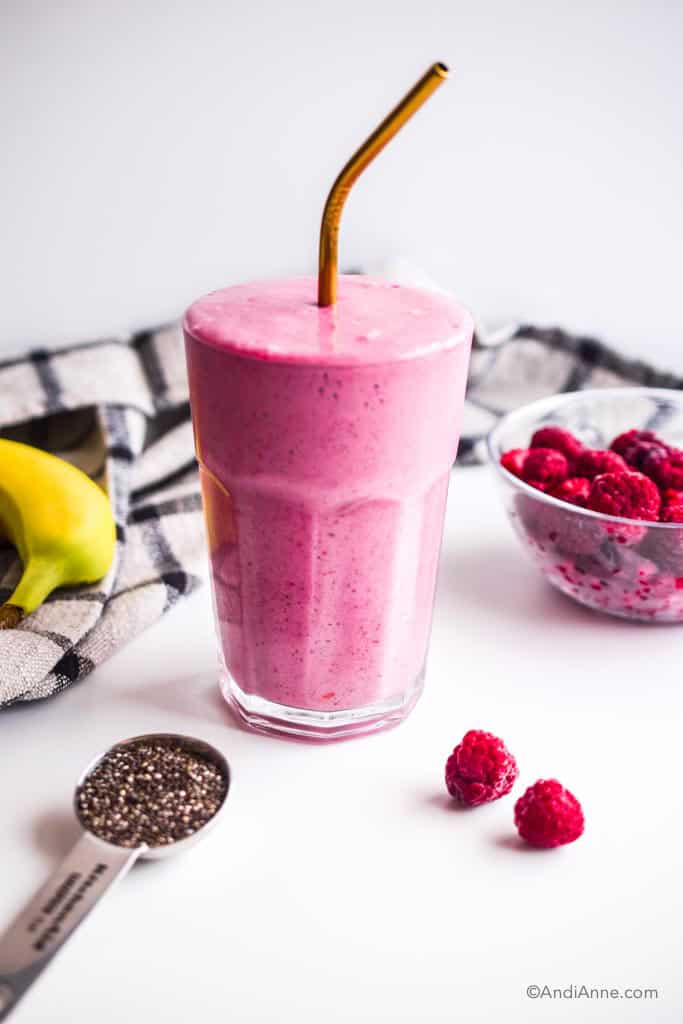 glass with raspberry yogurt smoothie inside and gold straw. Bowl of raspberries, a banana, spoon of chia seeds and a kitchen napkin surround the glass.