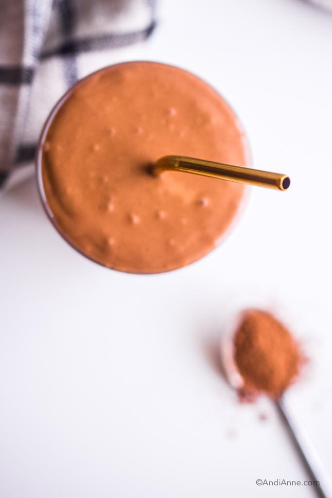 looking down on chocolate smoothie with gold straw. Spoon with cacao powder is blurred in the background.