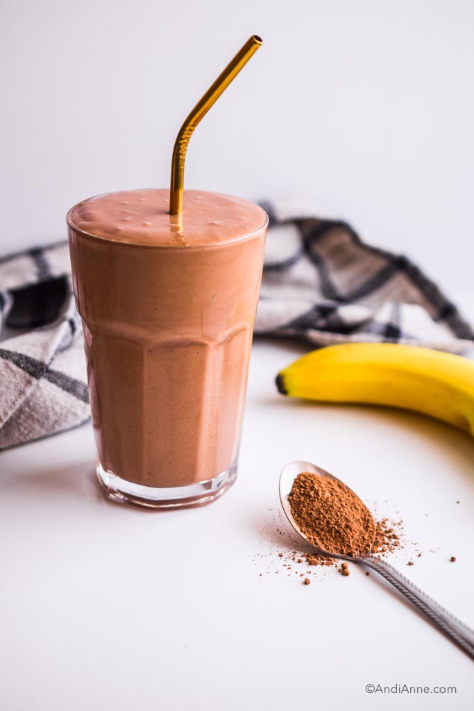 banana chocolate smoothie in a glass with gold straw. Kitchen napkin, banana and spoon of cacao powder surround it.