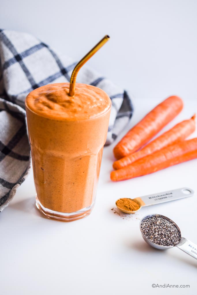 carrot cake smoothie in a glass with metal straw with 3 carrots, spoon of cinnamon and spoon of chia seeds beside it