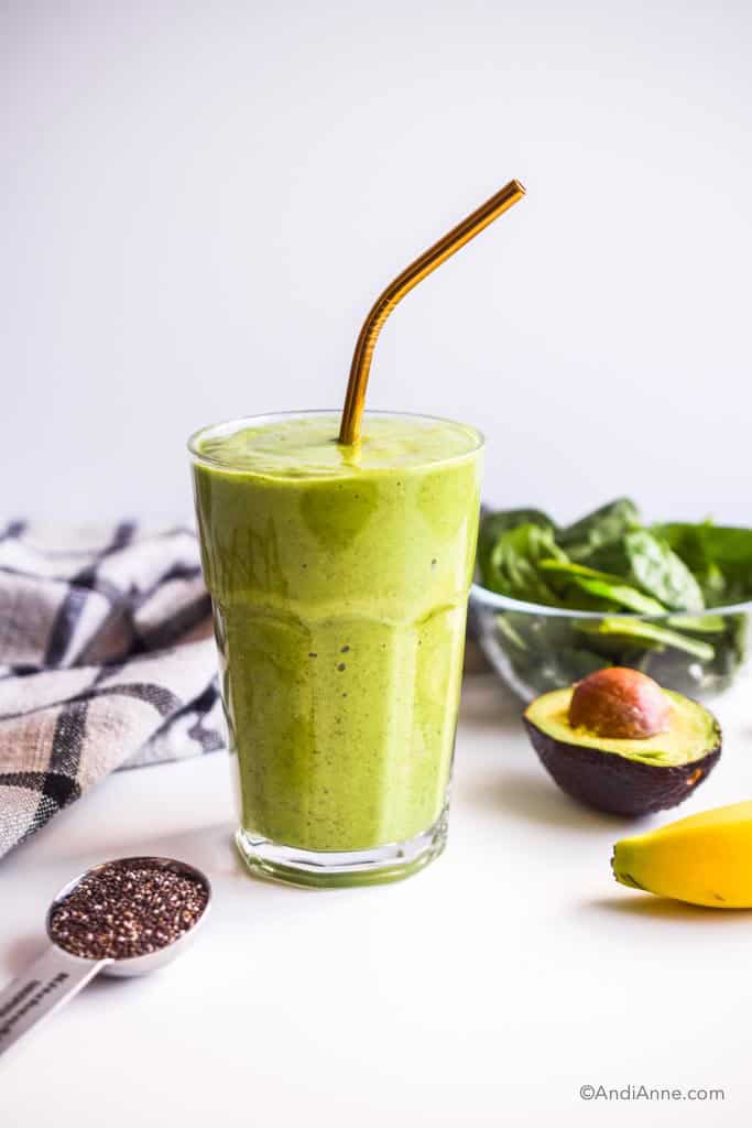green smoothie in a glass with spoon of chia seeds, half of an avocado, banana and bowl of spinach beside it