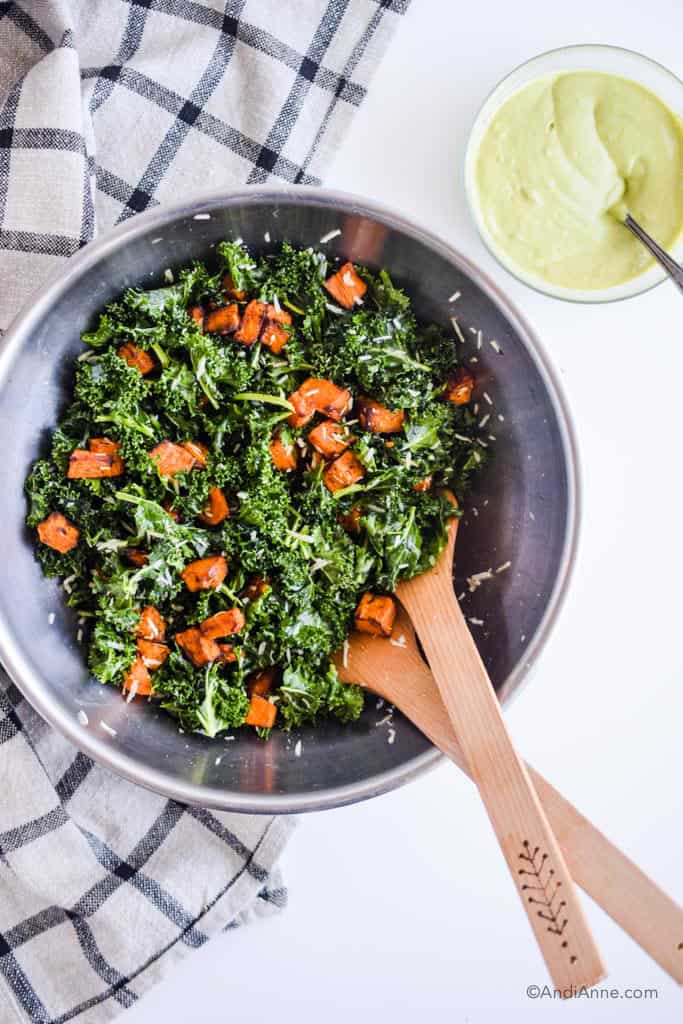 metal bowl with kale, sweet potato and grated parmesan cheese and wooden salad spoons. Bowl of avocado dressing beside it.