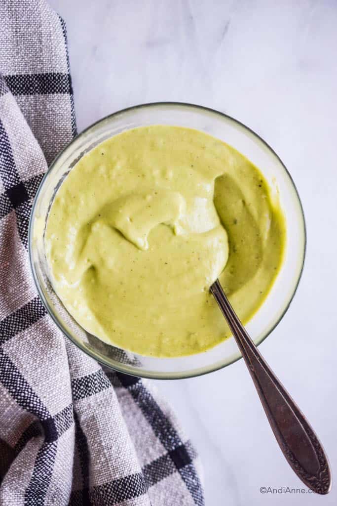 bowl of avocado dressing with a spoon and white and black kitchen towel beside it