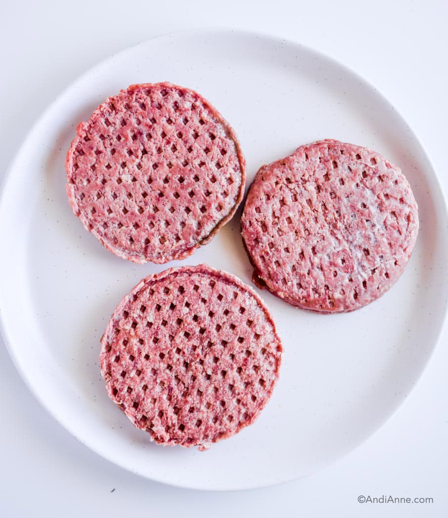 three raw hamburger patties on a white plate.