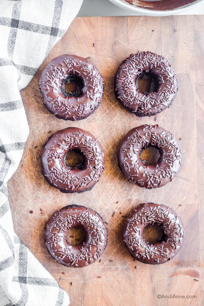 six chocolate baked donuts on cutting board with kitchen towel beside it.