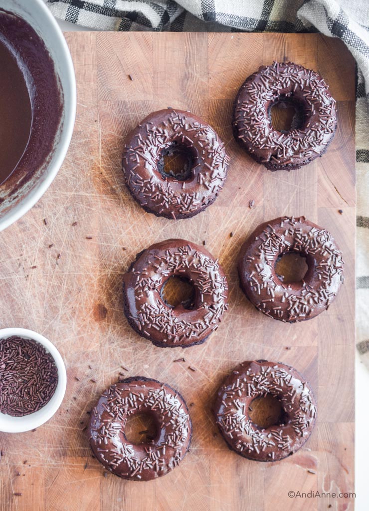 chocolate baked donuts with sprinkles on a cutting board with glaze in a bowl and chocolate sprinkles beside it