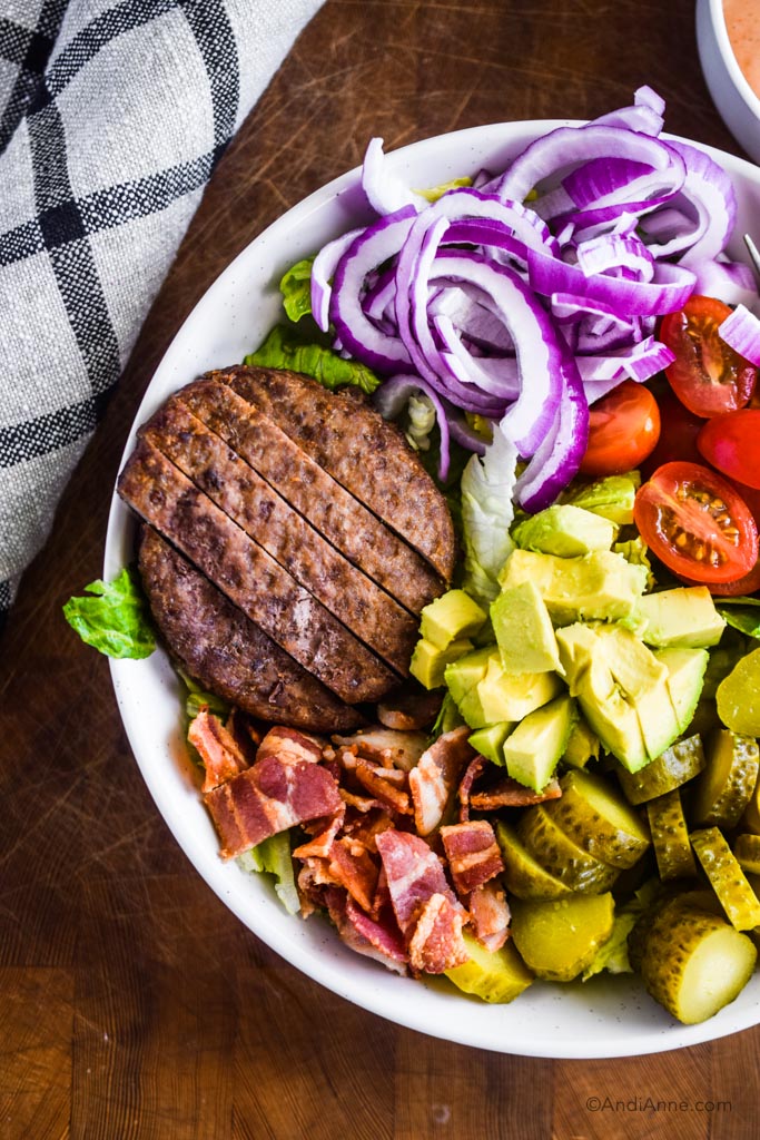 close up of burger salad bowl including burger patty, avocado slices, sliced red onion and bacon.