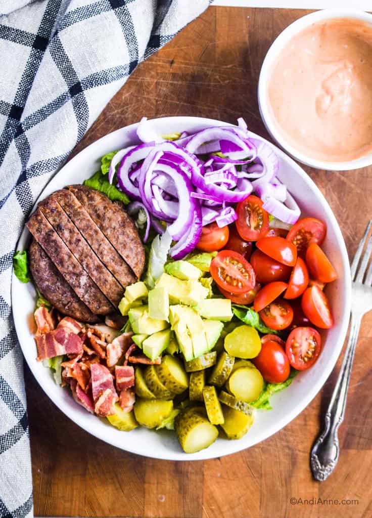 burger salad bowl on white plate with fork and kitchen towel beside it. Salad dressing in small bowl top right.
