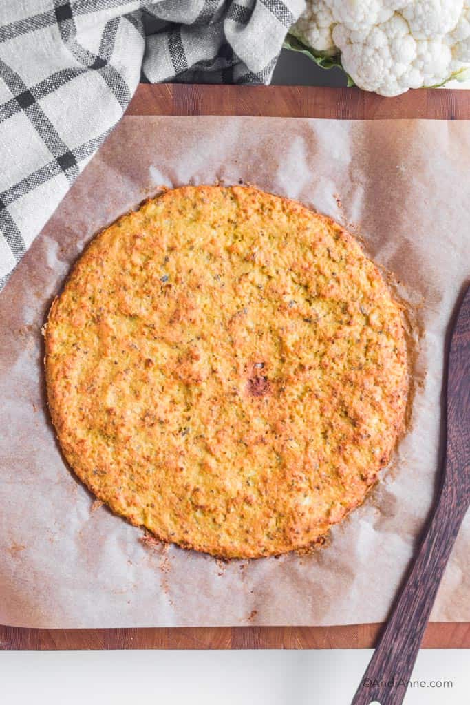 homemade cauliflower pizza crust on parchment paper with kitchen towel beside it and wood spatula