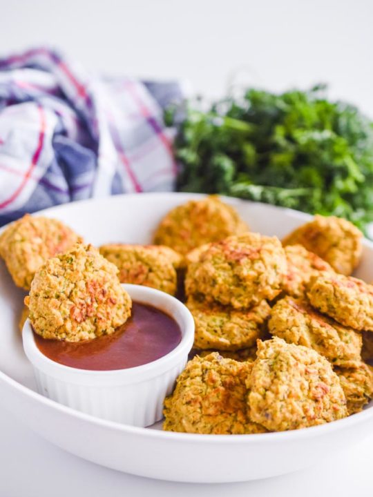 chickpea nuggets in a white bowl with a small cup of ketchup. Parsley and kitchen towel in the background.