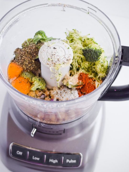All ingredients inside a food processor including grated zucchini, broccoli, Italian seasoning, eggs, spices and chickpeas.