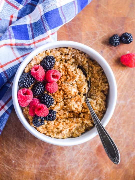 steel cut oats in a white bowl with fresh berries, cinnamon and a spoon. Blue plaid kitchen towel beside bowl.