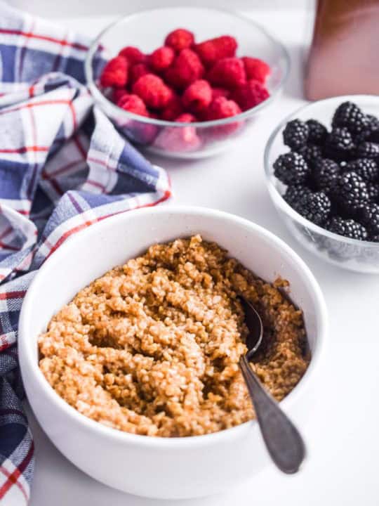 instant pot steel cut oats in a bowl with a spoon. Two glass bowls with raspberries and blackberries beside it along with plaid kitchen towel.