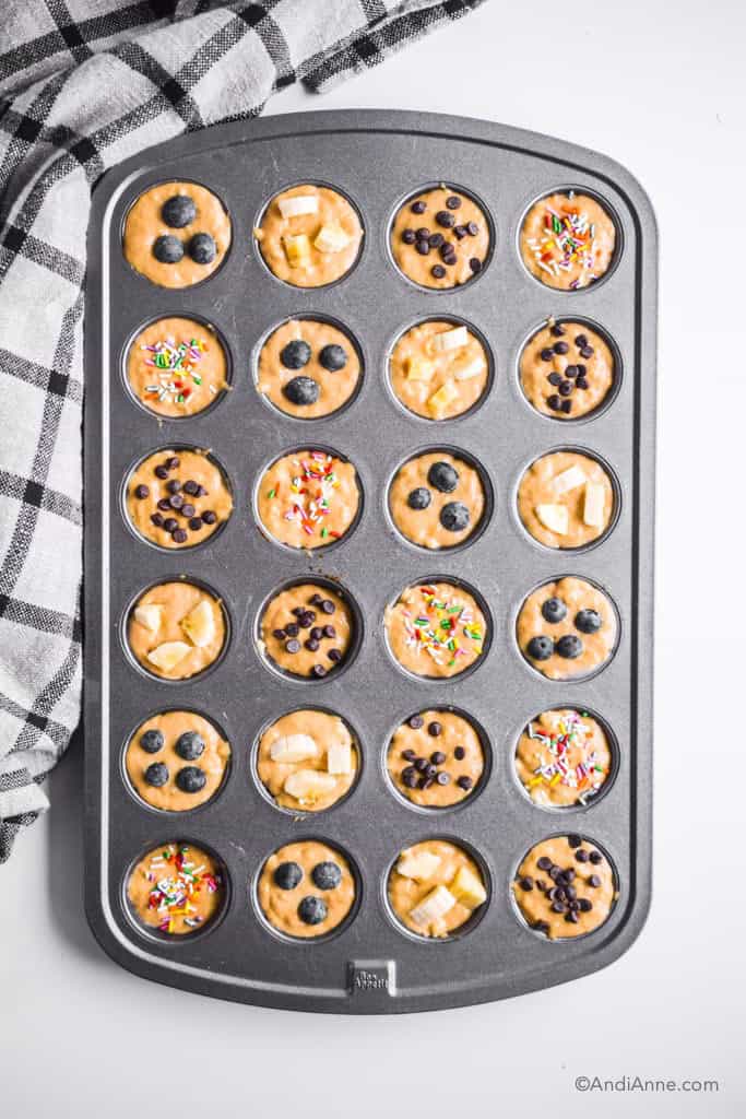 raw muffin batter inside metal mini muffin pan. berries, banana, chocolate and sprinkles top each one