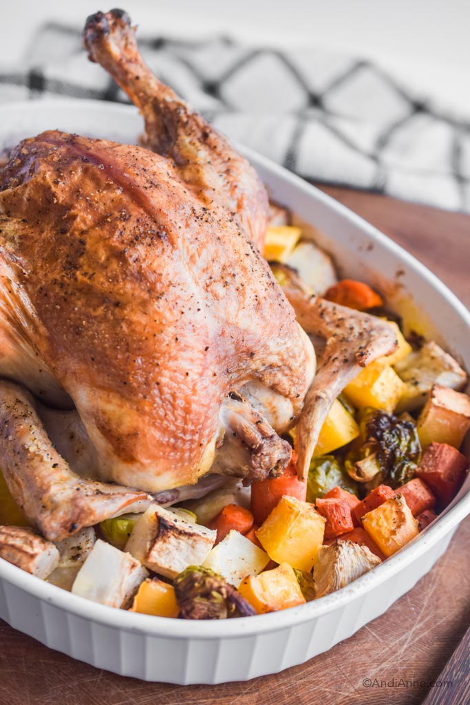 Roasted whole chicken in white dish surrounded by cooked root vegetables. On wood cutting board with kitchen towel in background.
