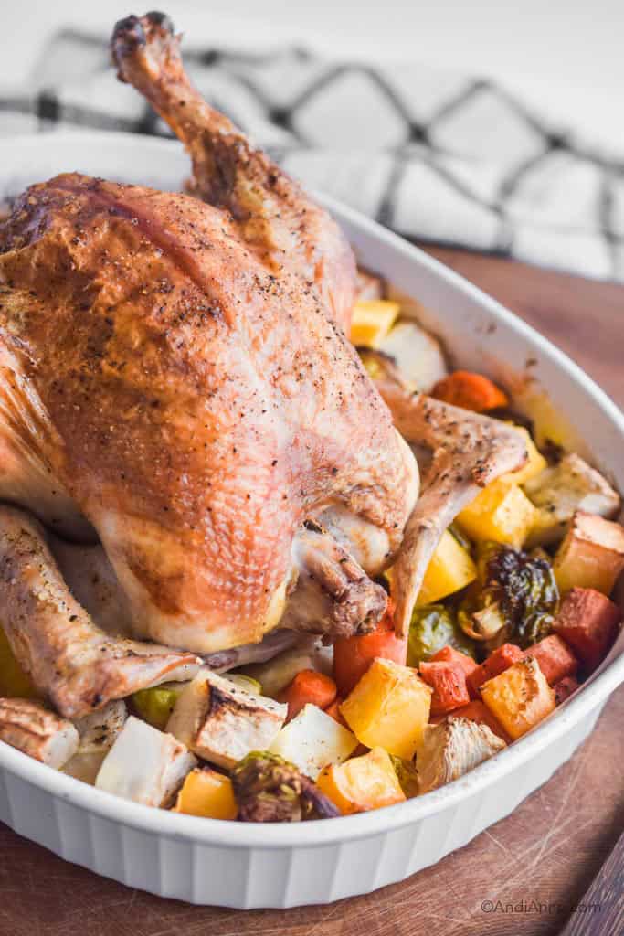 Roasted whole chicken in white dish surrounded by cooked root vegetables. On wood cutting board with kitchen towel in background.
