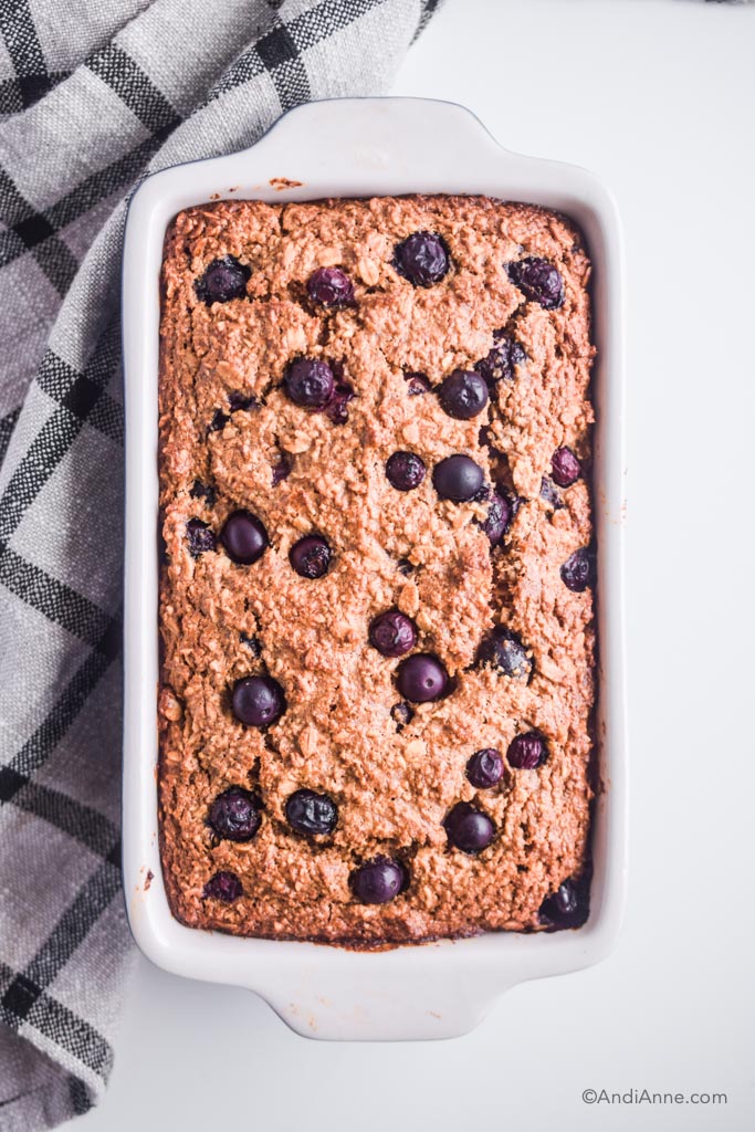 oatmeal blueberry breakfast loaf in a white ceramic dish with kitchen towel on the side.