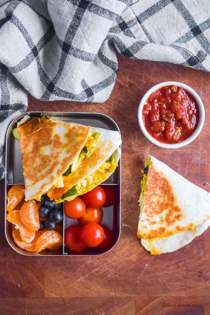 quesadilla in steel container with cherry tomatoes, oranges and grapes. Cup of salsa beside it