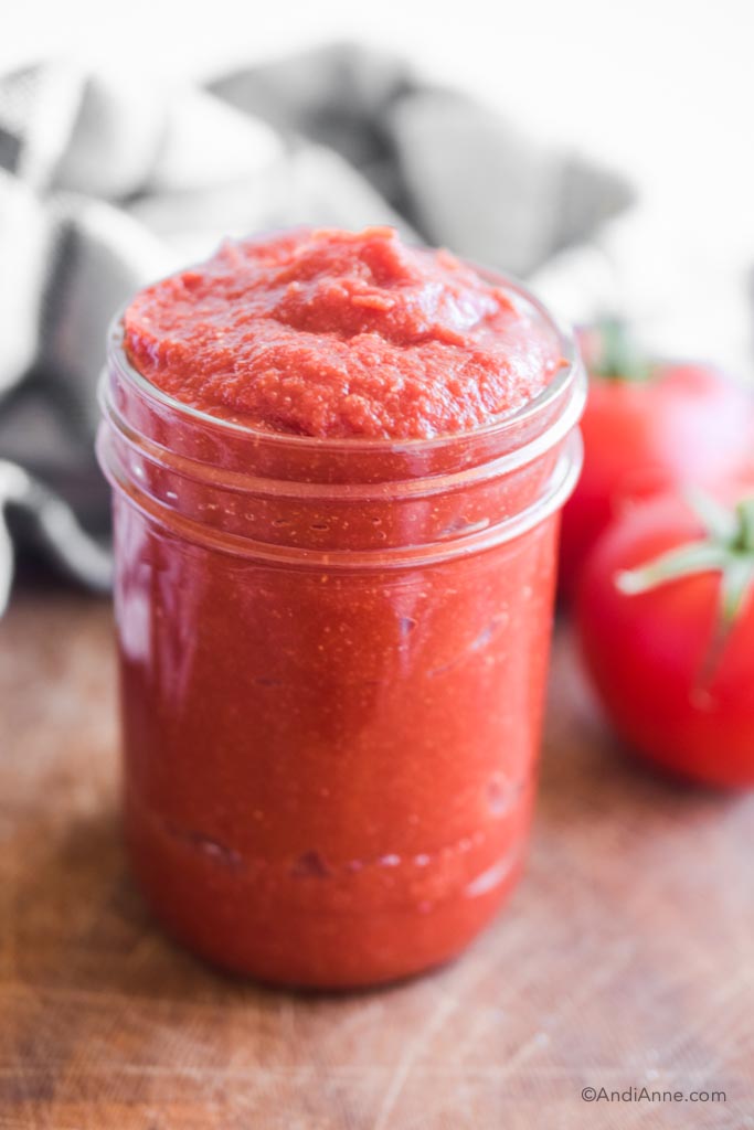 homemade ketchup in a mason jar with tomatoes and kitchen towel blurred in the background