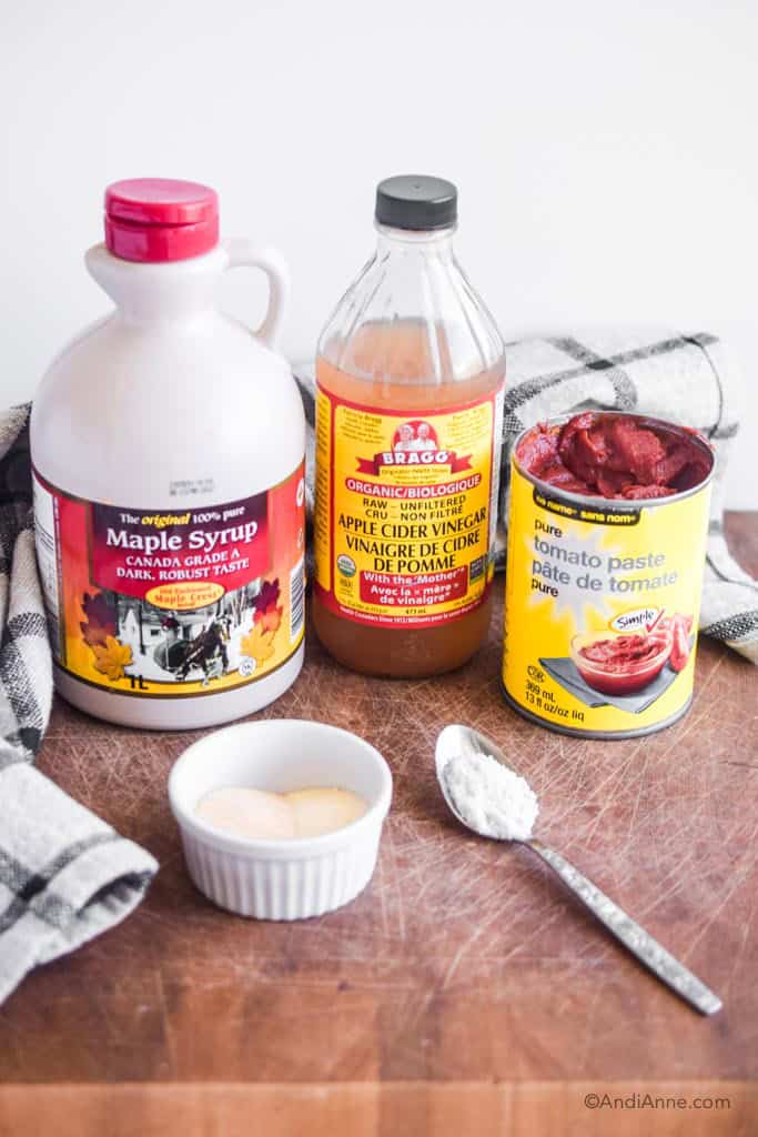 ingredients to make homemade ketchup on a wood block. Maple syrup, apple cider vinegar, canned tomato paste, garlic powder and onion powder in a white container, and sea salt on a spoon.
