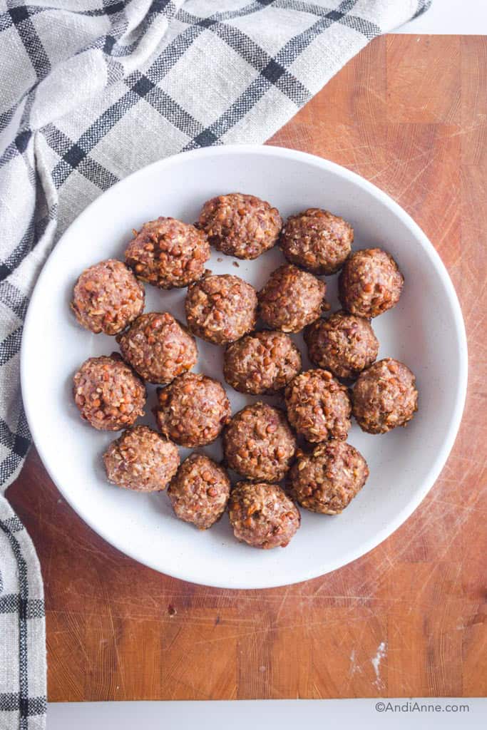 White bowl with cooked meatballs in it on wood cutting board with kitchen towel beside it