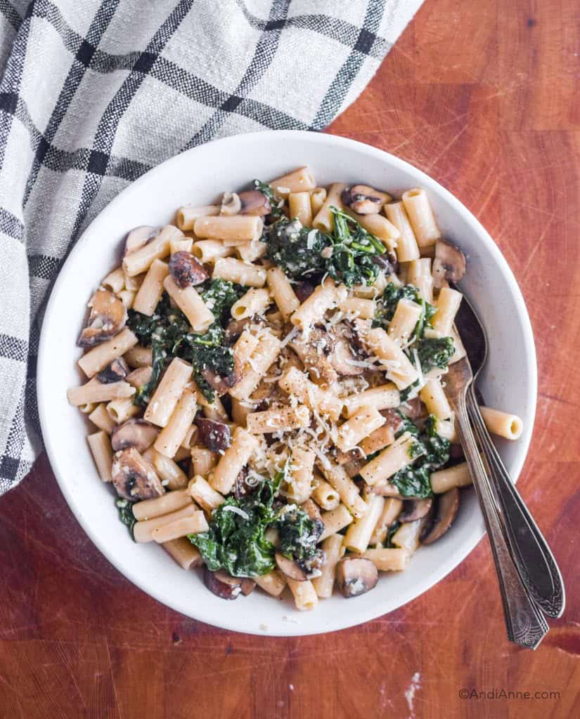brown rice penne pasta with parmesan, kale and mushrooms in a white bowl with silver spoon and fork on the side.