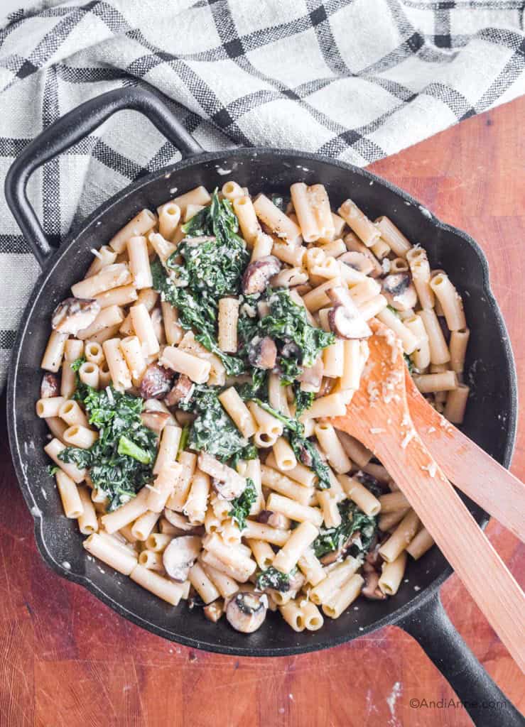 penne pasta with kale, mushrooms and parmesan cheese in a cast iron skillet and two wooden spoons. Pan on a wood board with kitchen towel in corner.