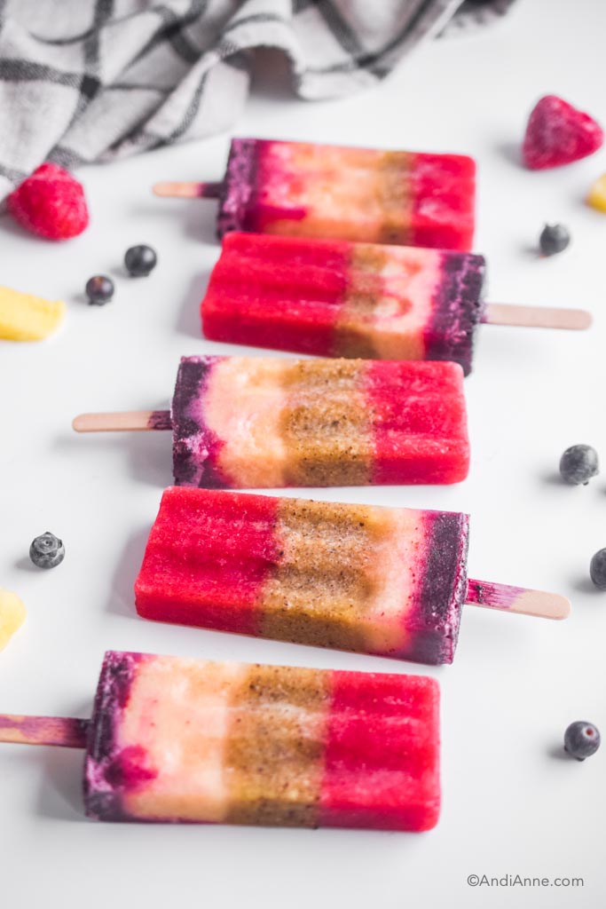 a row of fruit popsicles on a white counter with frozen fruit around them