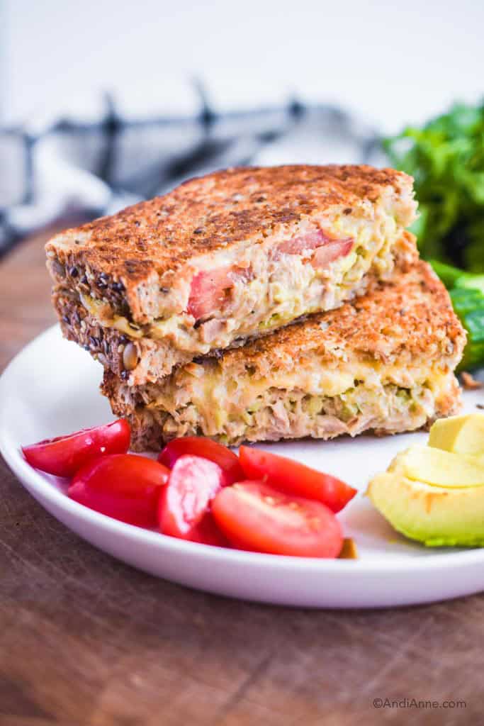 avocado tuna melt on a white plate with sliced grape tomatoes, avocado and cucumber beside it.