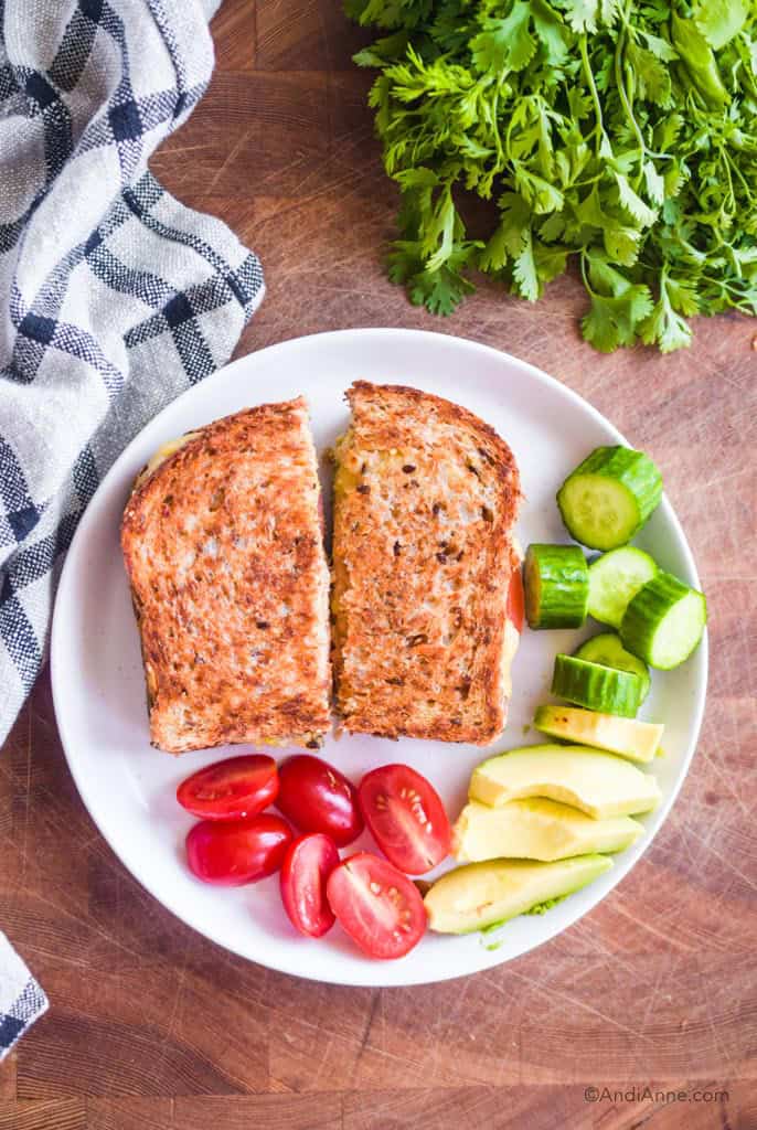 avocado tuna melt sliced in half on a white plate with sliced grape tomatoes, avocado and cucumbers. The plate is on a cutting board with kitchen towel and parsley surrounding it.