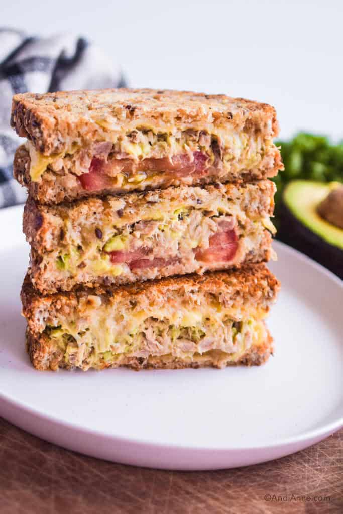 three slices of avocado tuna melt stacked on top of eachother on a white plate. Avocado and parsley are in background.