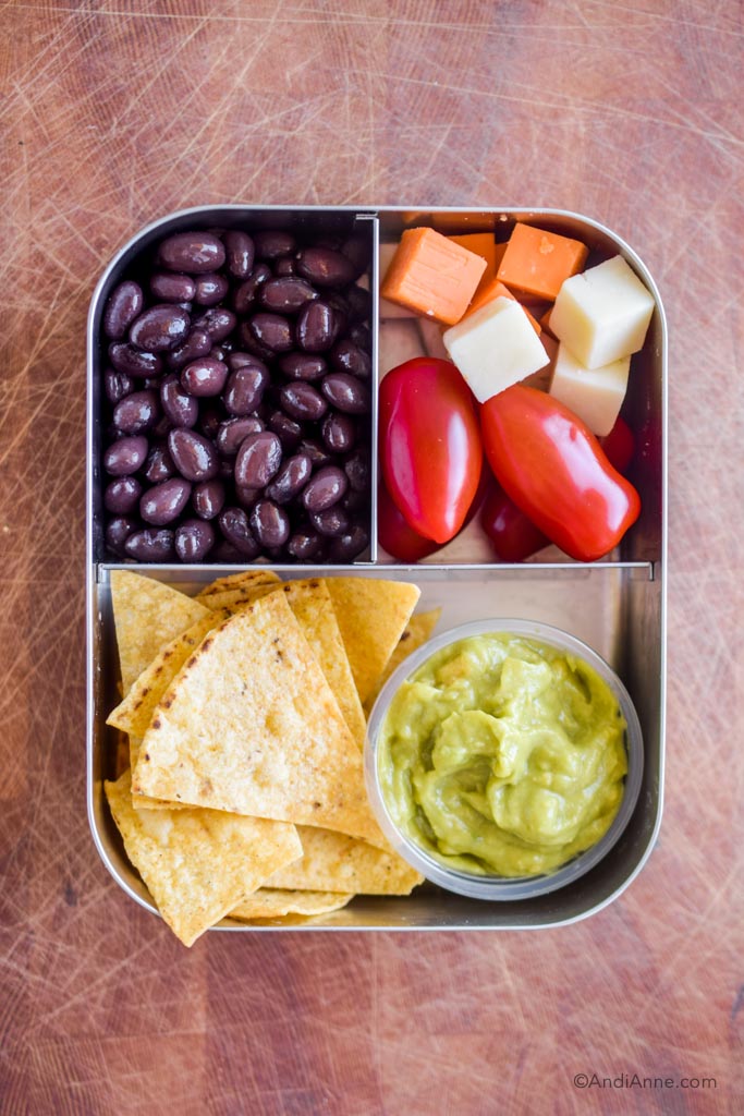 Stainless steel bento box lunch with black beans, grape tomatoes, cheese cubes, tortilla chips and small container of guacamole.