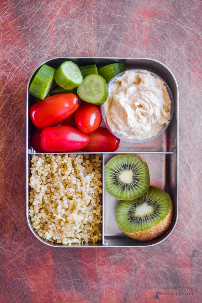 Stainless steel bento box lunch with quinoa, kiwi slice, grape tomatoes, cucumber slices, and hummus.