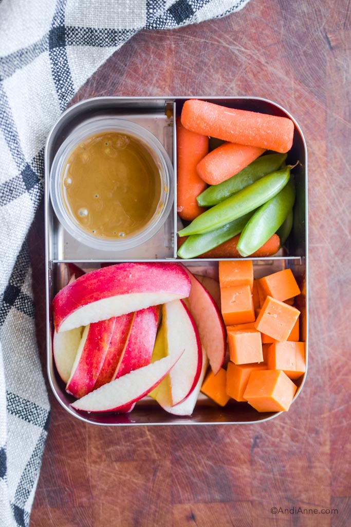 Stainless steel bento box lunch with apple slices, cheese cubes, carrots, snap peas, and a small container of seed butter.