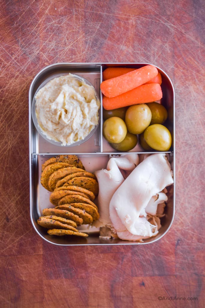 Stainless steel bento box lunch with deli turkey slices, handful of crackers, baby carrots, olives and small container of hummus.