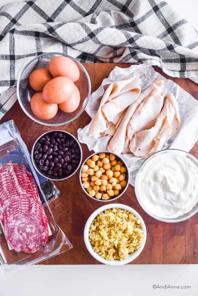 Hard boiled eggs, deli meat, beans, greek yogurt, quinoa and salami all in separate bowls on a wood cutting board.