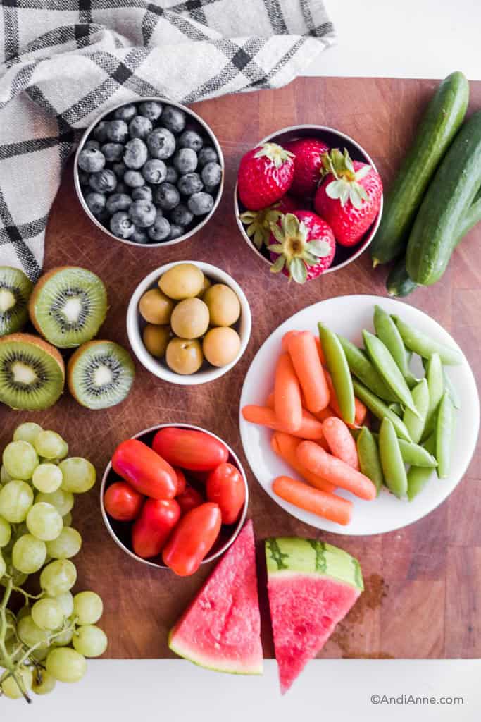 Berries, olives, sliced kiwi, snap peas, carrots, grape tomatoes, watermelon, and grapes in bowls and on a wood cutting board.