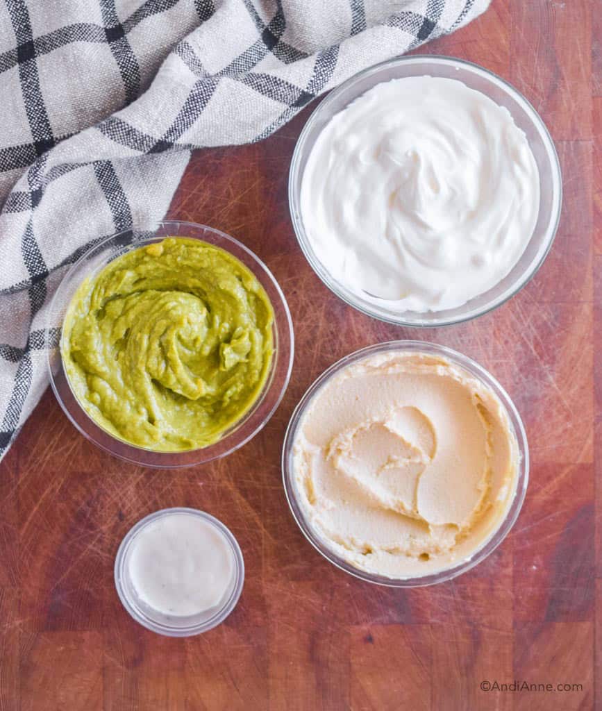 four bowls on a wood cutting board. One with guacamole, greek yogurt, hummus and ranch dip.