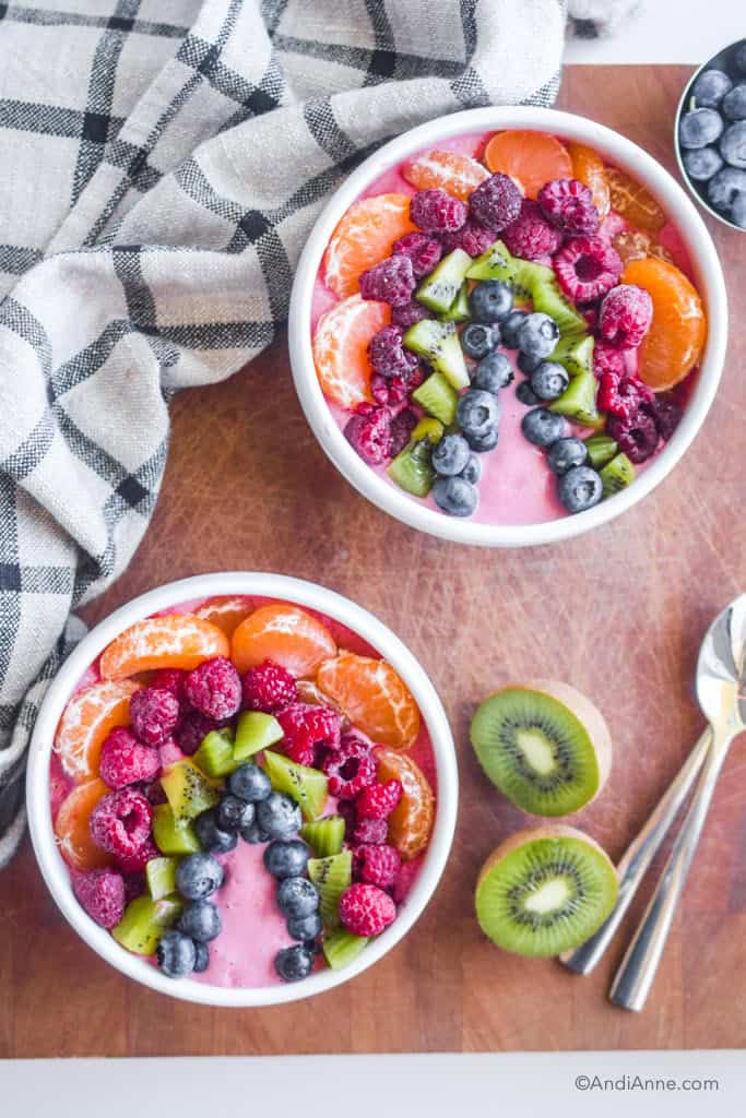 two rainbow smoothie bowls with fruit toppings on a wood cutting board. Kitchen towel, sliced kiwi and two spoons surround the bowls.