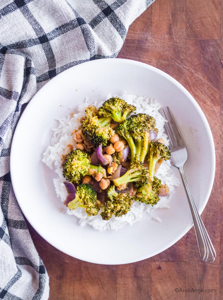 rice and broccoli chickpea stir fry in a white dish with a fork. Dish is on a wood cutting board with kitchen towel beside it. 