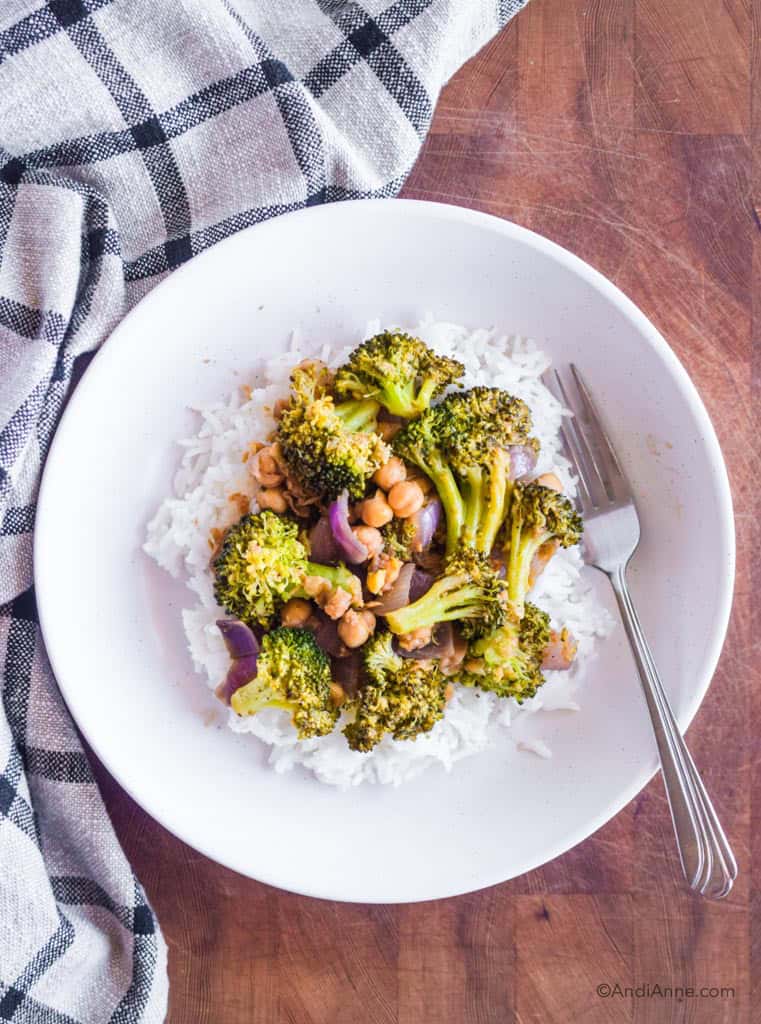 rice and broccoli chickpea stir fry in a white dish with a fork. Dish is on a wood cutting board with kitchen towel beside it.
